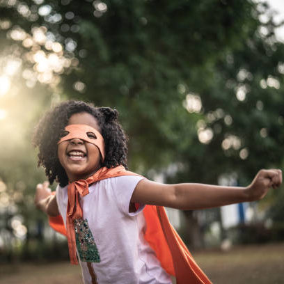 Joyful young black girl smiles at the camera wearing a superhero cape and mask.